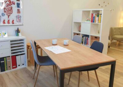 Besprechungsecke. Tisch mit 3 Stühlen. Bücherregal im Hintergrund. Arbeitsbereich in der Praxis für ganzheitliche Ernährungsberatung, ernährungs-psychologische Beratung und Hypnosetherapie in Dübendorf, Stettbach.